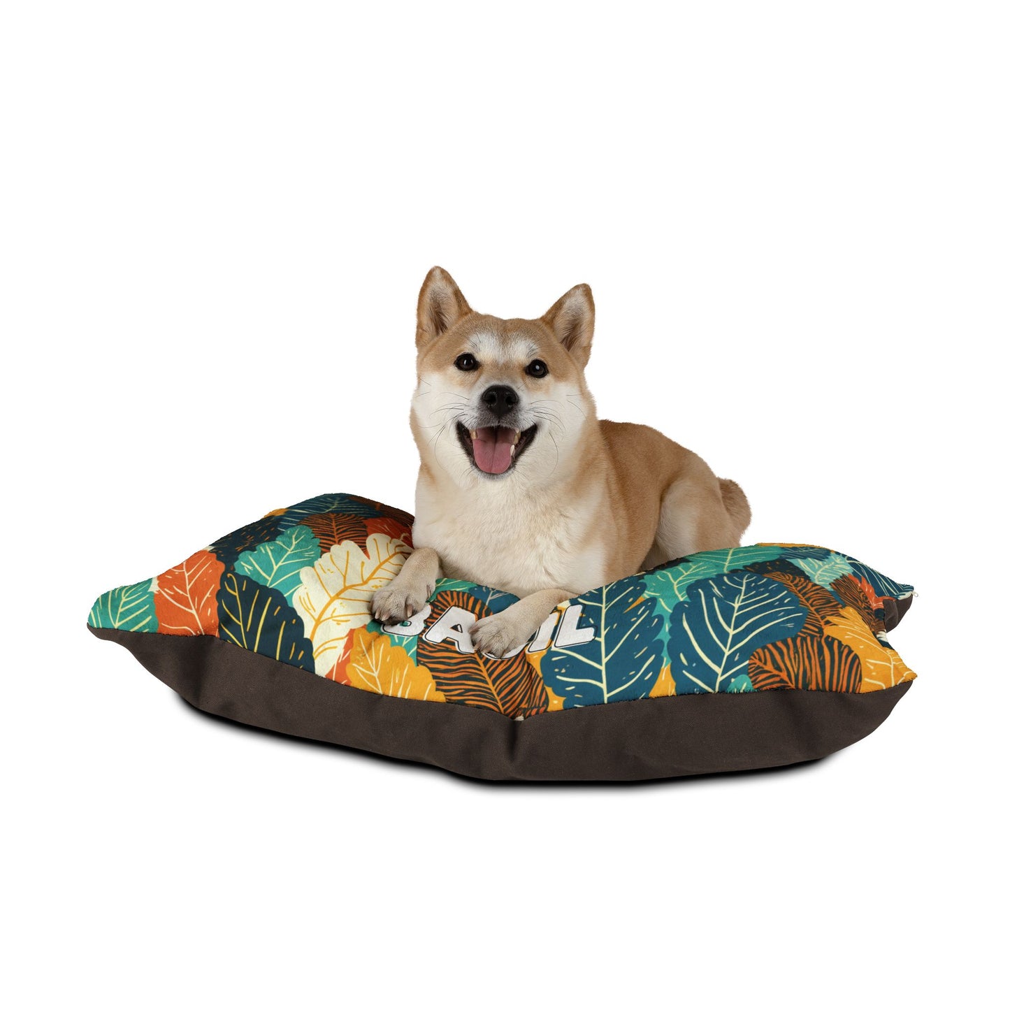 A happy Shiba Inu dog with tan and white fur lies on a colorful cushion decorated with a vibrant leaf pattern and dark brown edges, offering luxurious comfort and durable construction against a plain white background. The dogs mouth is open, tongue out, ears perked up.
