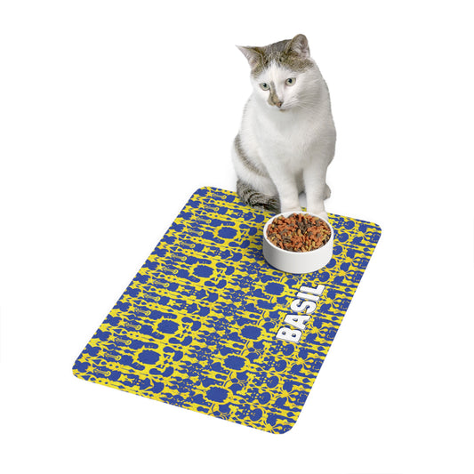 A white and gray cat sits beside a food bowl filled with kibble, placed on an African-inspired blue and yellow patterned mat. The mat has the name BASIL printed in bold white letters. The background is plain white.