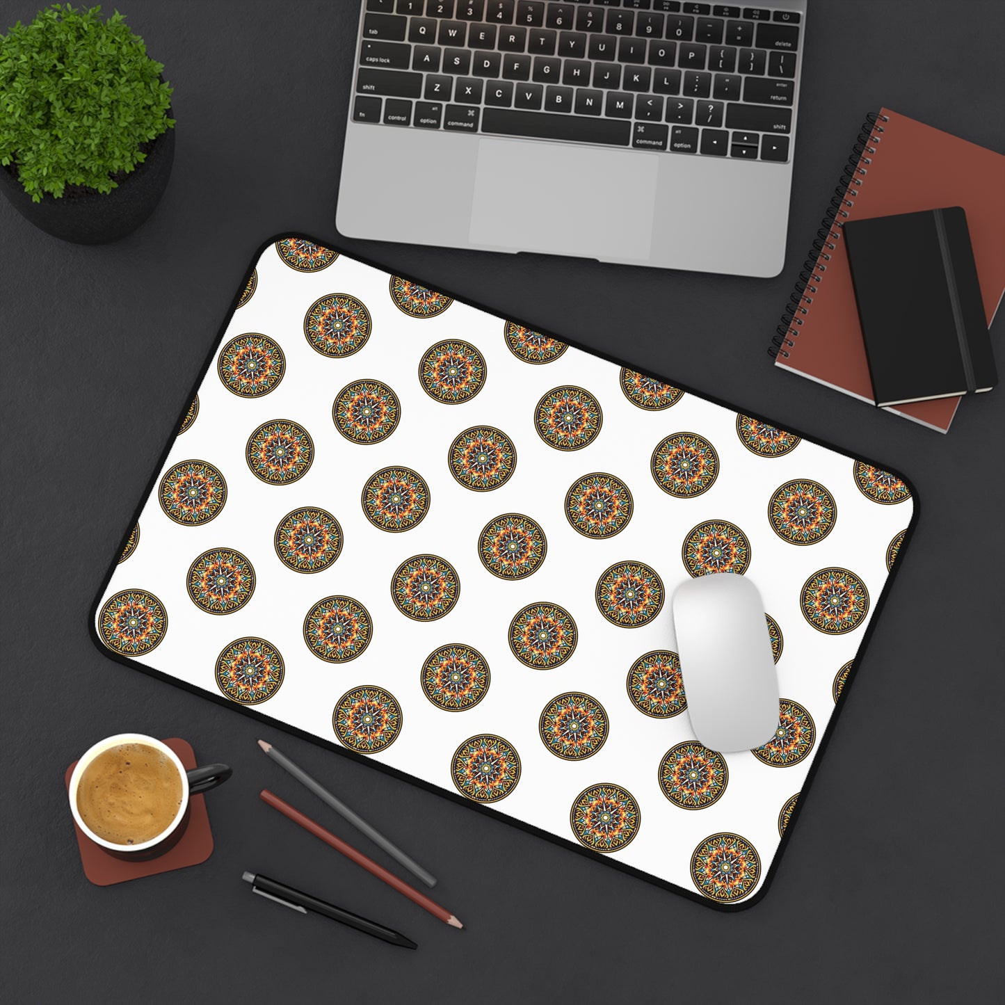 A neatly arranged black desk with a laptop, small potted plant, notebooks, pencils, coffee, and a white mouse on an ergonomic neoprene desk mat featuring a repeating colorful mandala pattern—a vibrant workspace accessory.