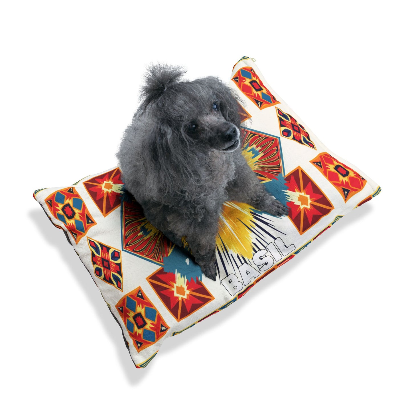 A gray poodle with curly fur sits on a colorful rectangular dog bed decorated with African-inspired geometric red, yellow, blue, and orange patterns. The name “BASIL” is printed in large white letters on one corner of the bed. The background is white.