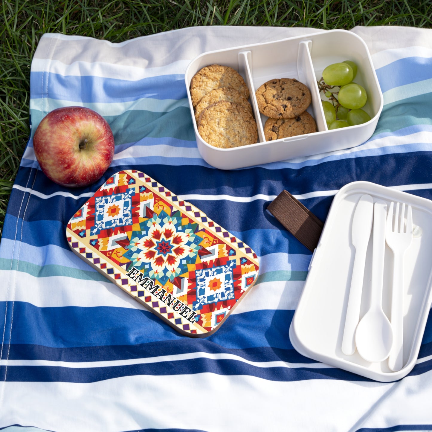 A colorful African-inspired bento lunch box labeled EMMANUEL sits on a striped blue picnic blanket. Next to it are a red apple, green grapes, cookies in a divided container, and white plastic utensils in an open case on green grass.