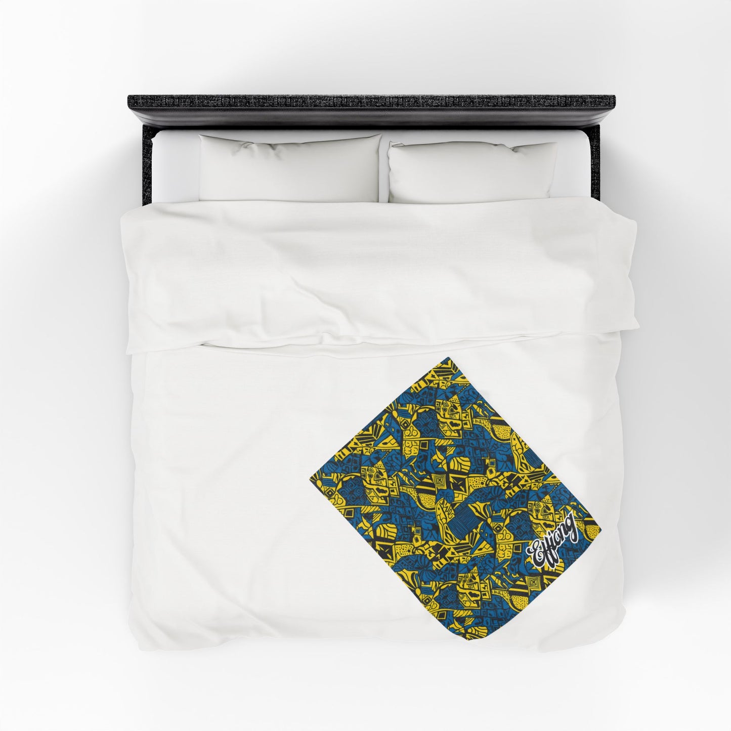 A neatly made bed with white bedding and two pillows. In the lower right corner, an African-inspired blanket with a yellow abstract pattern and white script text is partially folded, adding a vibrant pop of color to the plain bedding.