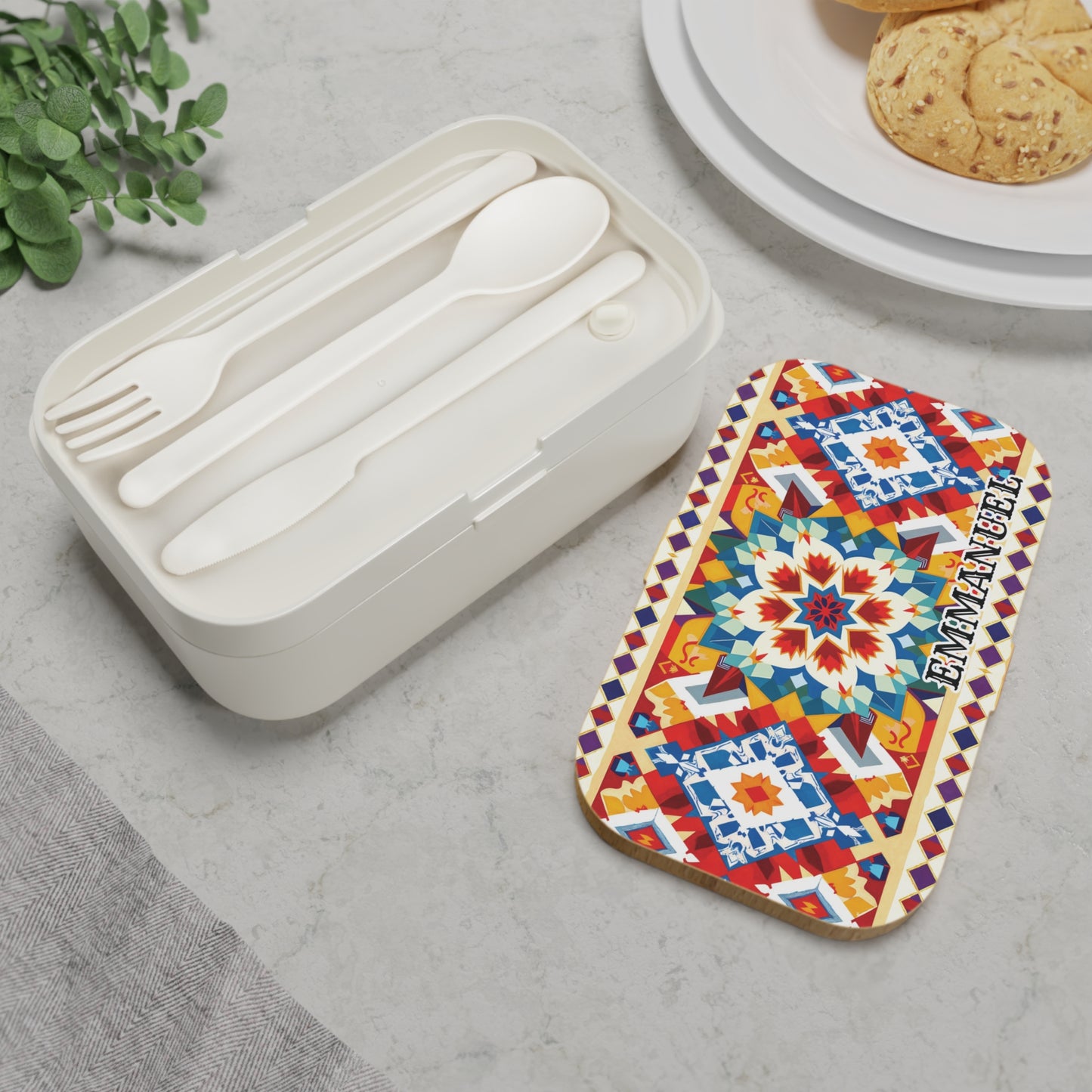 A personalized, African-inspired bento lunch box with a colorful geometric lid labeled EMMANUELLE sits on marble. Inside the BPA-free lunchbox are a white fork, spoon, and knife. Nearby are a plant, gray napkin, and cookies on a plate.