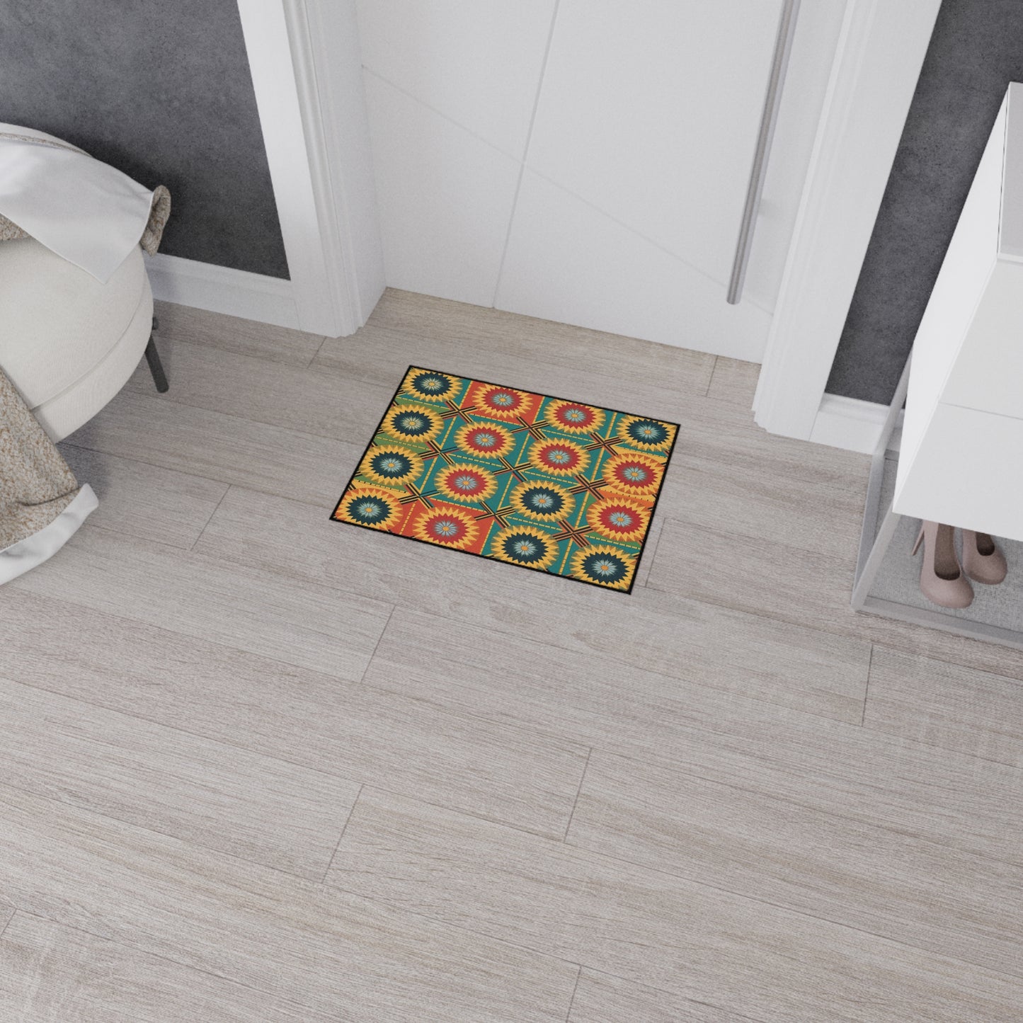 A small, colorful mat with African-inspired aesthetics and a geometric sunburst pattern in red, yellow, green, and blue sits on a light wood floor near a white door. Nearby are a white round chair, gray walls, a white cabinet, and beige high-heeled shoes on a shelf.