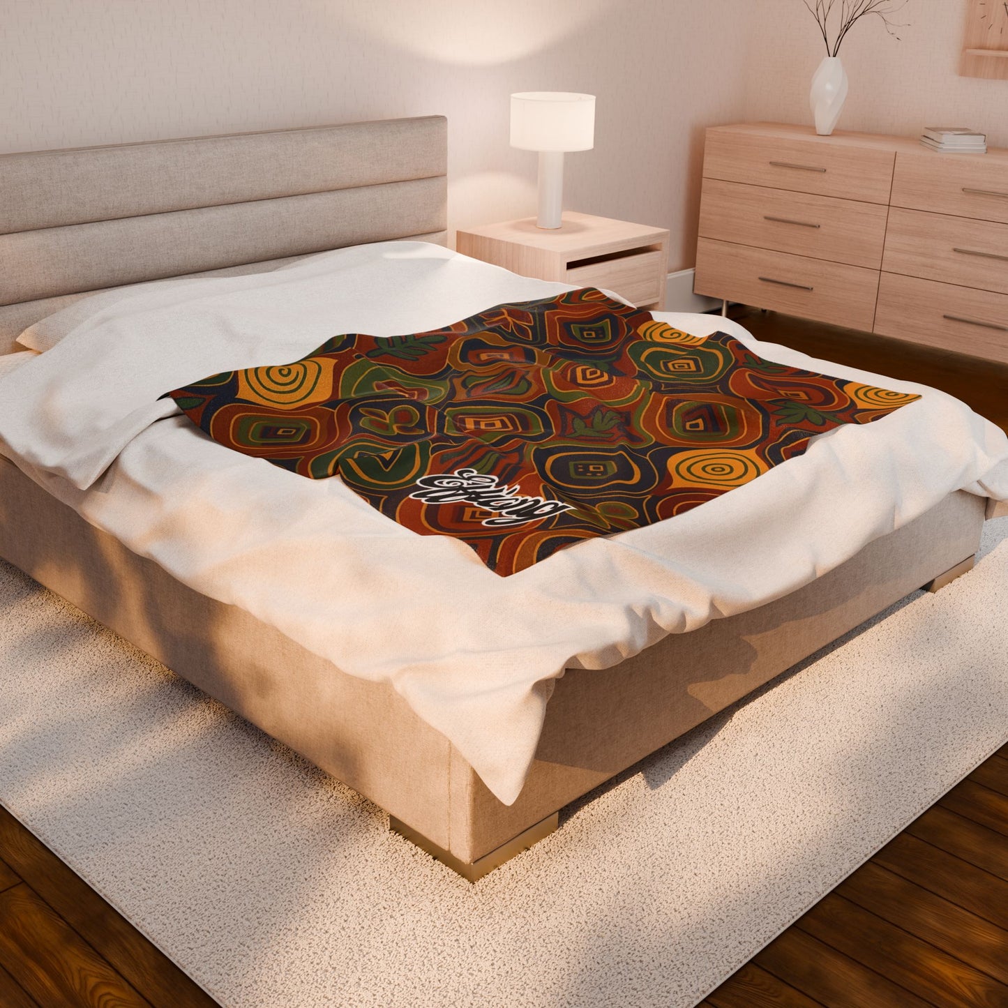 A neatly made bed in a modern, softly lit bedroom features an African-inspired personalized blanket with colorful swirls and geometric shapes laid across the foot. The word “Ebony” appears on this unique keepsake gift, complementing the light wood furniture and white rug.