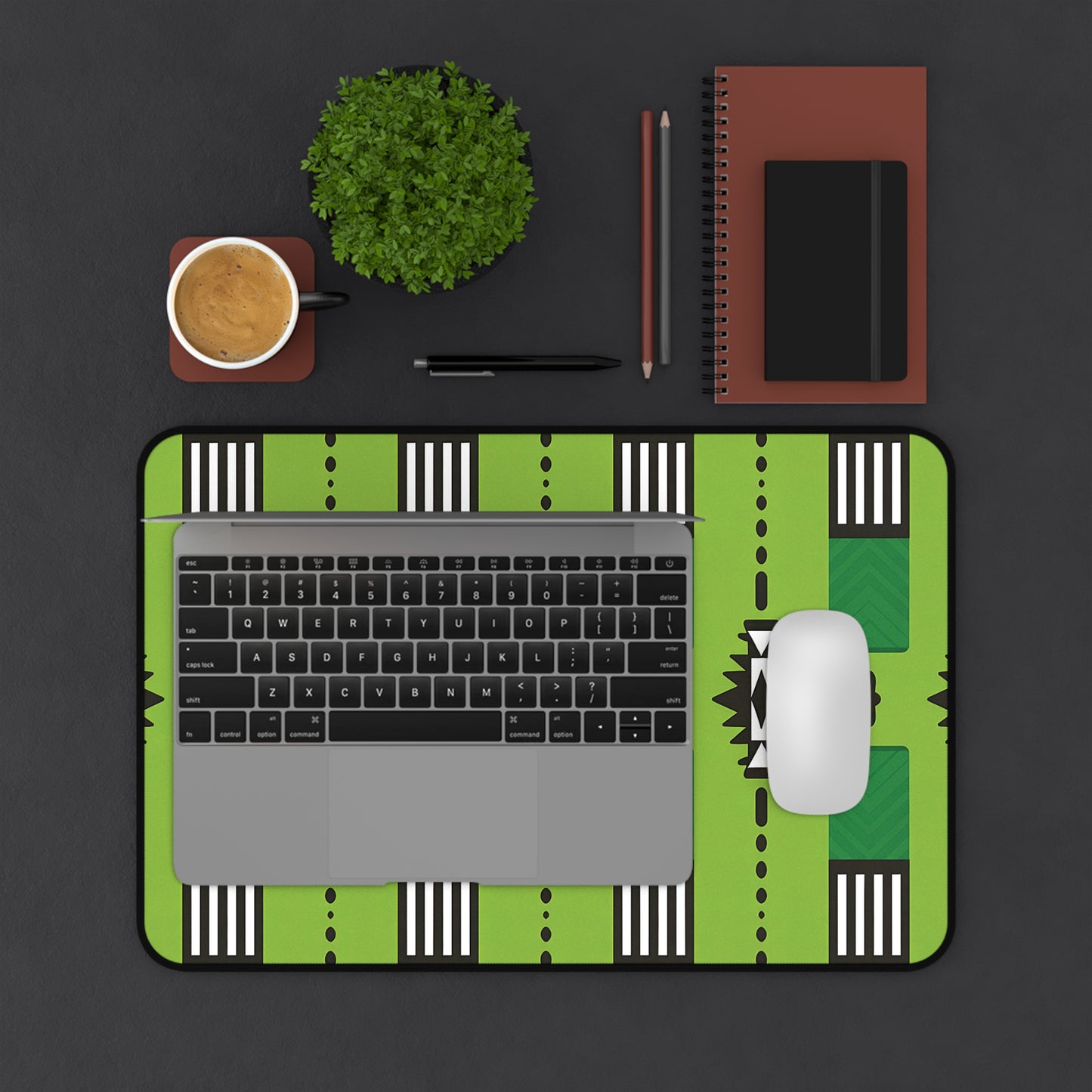 A top-down view of a workspace with a laptop and white mouse on a green patterned neoprene desk mat, a coffee cup, small potted plant, two pencils, pen, and stacked brown notebooks with a black stripe, all on a dark surface.