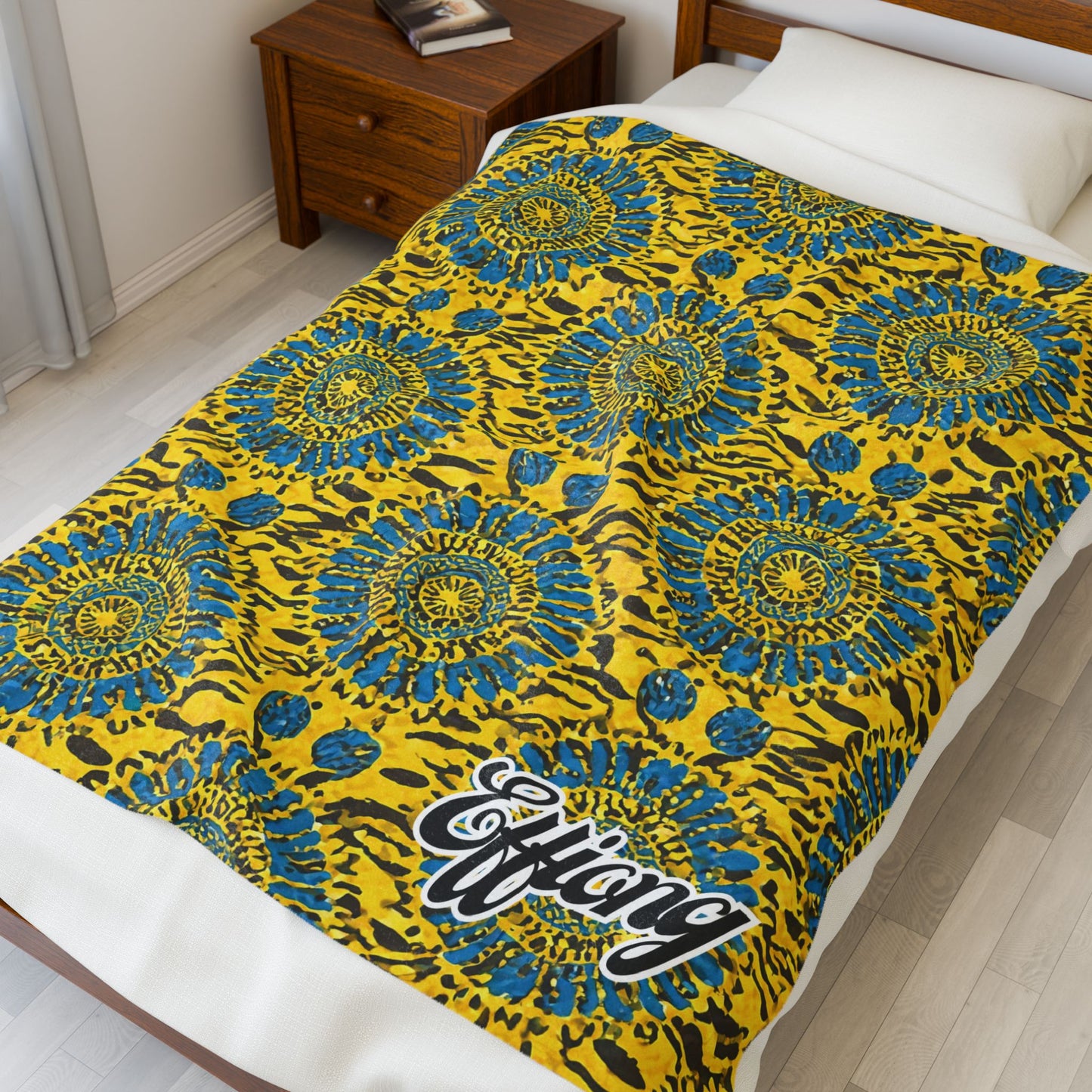 A bed with a bright yellow and blue African-inspired blanket featuring circular sunburst designs, black accents, and the word “Efiang” in bold, white script. The bed is next to a wooden nightstand in a white, minimal bedroom.