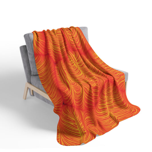 A gray, modern armchair with light wooden legs displays an African-inspired fleece blanket featuring a vibrant orange and yellow swirling pattern. The bold design stands out, adding cozy home decor and warmth to the neutral chair.