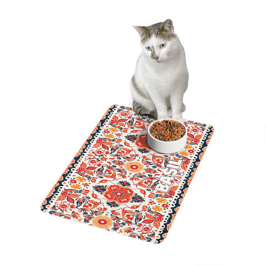 A white and gray cat sits on an African-inspired decorative mat with red and orange floral patterns, next to a bowl of cat food. The word BASIL is printed on the mat. The background is plain white.