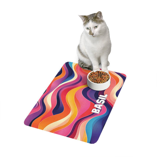 A white and grey cat sits behind a bowl of dry cat food on a colorful, African-inspired wavy-patterned mat. The mat features bright swirls of orange, pink, blue, and yellow with the name BASIL printed in bold white letters. The background is white.
