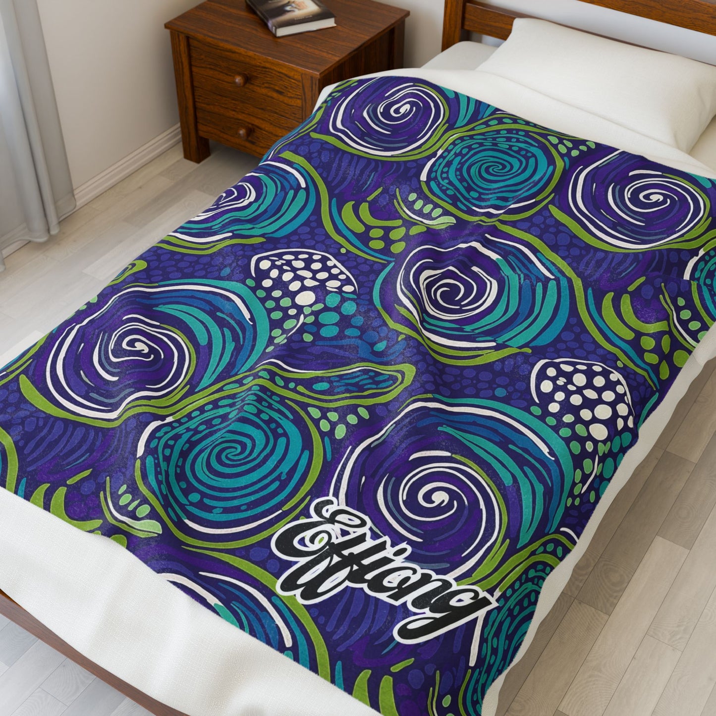 A bed with a colorful African-inspired blanket featuring swirling blue, purple, and green patterns accented by white dotted clusters. The custom gift is draped neatly, with Efflang in white script at the corner. A wooden nightstand sits beside the bed.