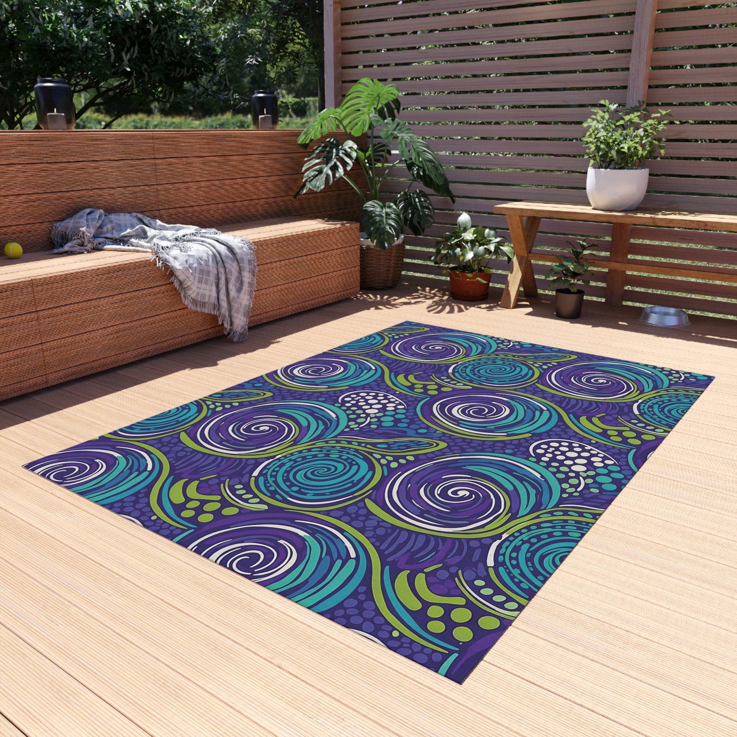 A modern outdoor patio with light wooden decking features an African-inspired outdoor rug with swirling blue, turquoise, and purple patterns. A wicker sofa with gray blanket, wooden table with plants, and lush greenery complete the cozy, sunlit space.