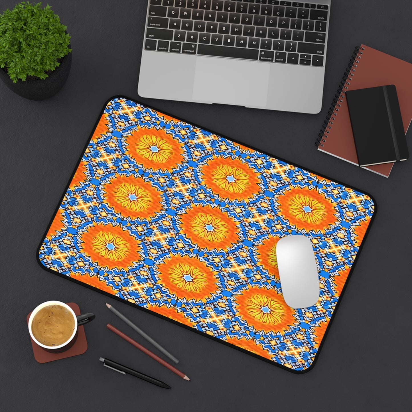 A large neoprene desk mat with a bright, geometric yellow, orange, and blue pattern adds flair to this colorful workspace accessory. A white mouse sits on it beside a laptop, notebooks, coffee cup, pencils, and a small potted plant.