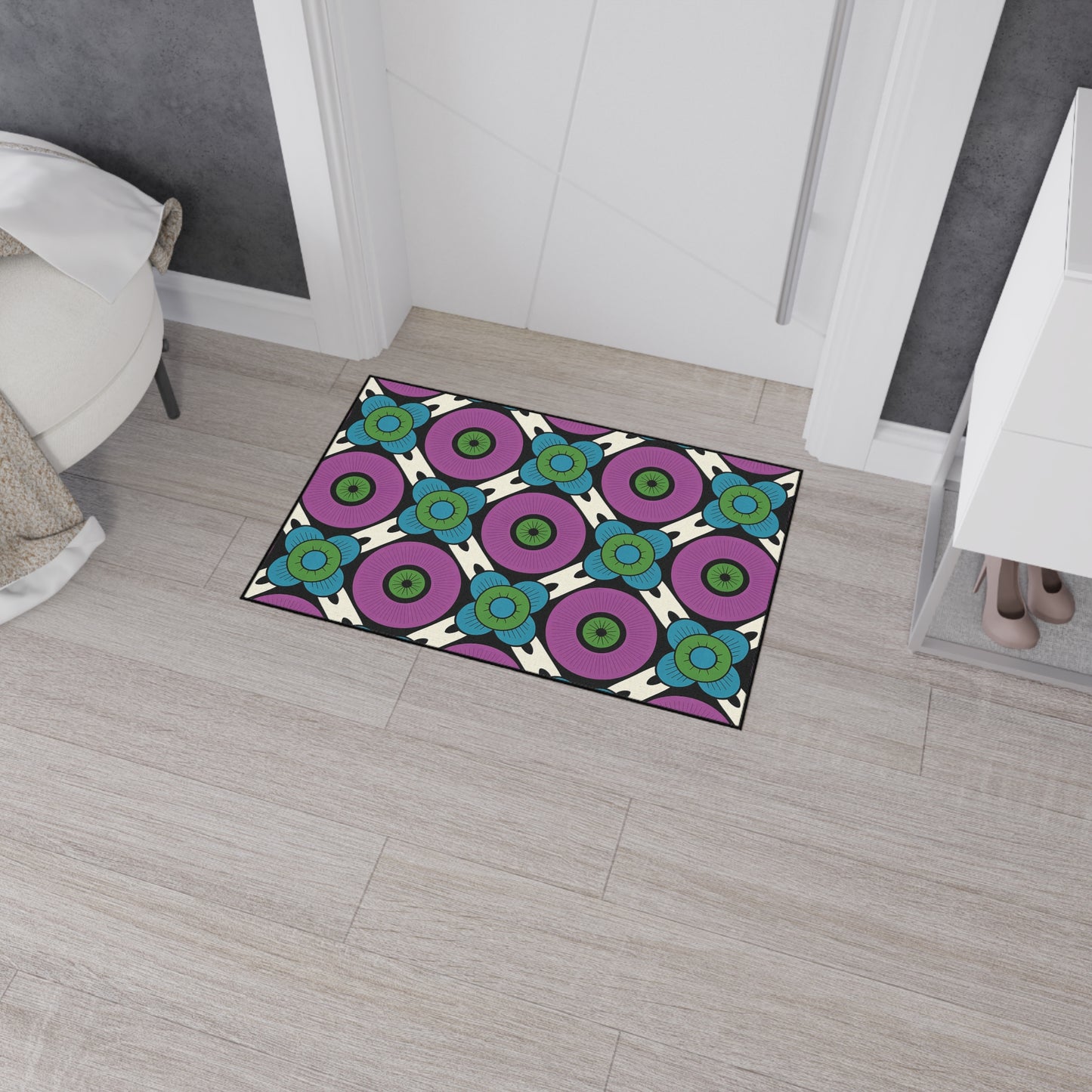 A geometric-patterned, non-slip doormat with purple, blue, green, and white circular designs lies on light wood flooring near a white cabinet and ottoman in a modern, gray-walled entryway. Shoes are visible on the bottom shelf of the cabinet.