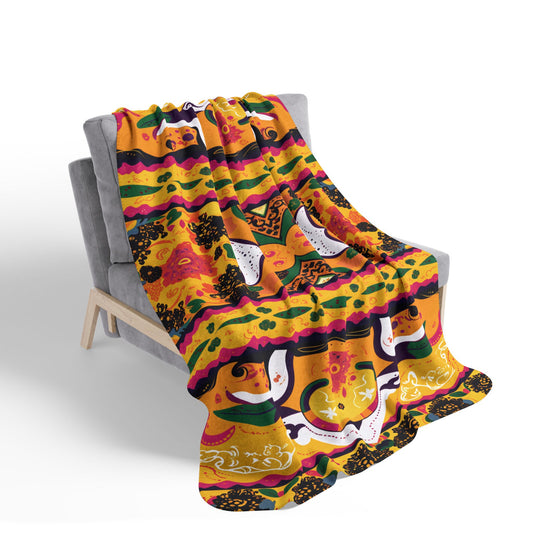 A soft gray armchair holds a colorful blanket draped over it, adding cozy home decor. The sherpa fleece blanket features a vibrant, Mexican-inspired pattern with bright florals, stripes, and sugar skulls. The background is plain white.