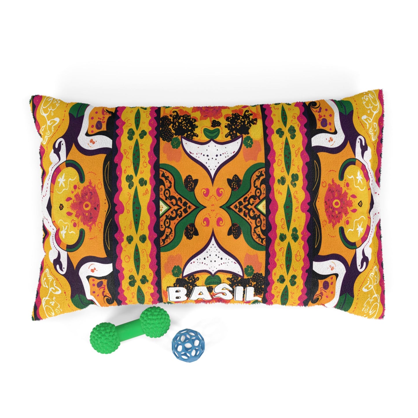 A rectangular pillow with BASIL embroidered at the bottom, featuring a vibrant, intricate African-inspired pattern in orange, yellow, green, and black. Below the pillow are two dog toys: a green bone-shaped toy and a blue ball with holes.