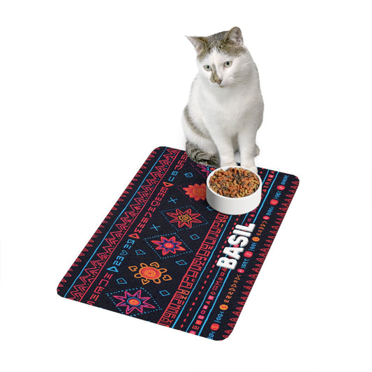 A white and gray cat sits beside a bowl of kibble on a rectangular mat with African-inspired geometric and floral patterns. The mat displays the name BASIL in bold white letters against a dark background, set on a plain white backdrop.