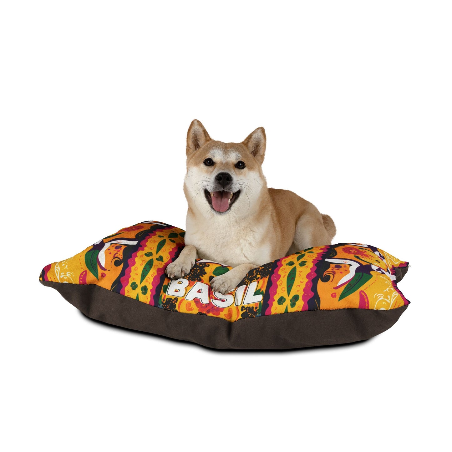 A happy Shiba Inu dog with tan and white fur lies on a colorful, African-inspired dog bed. The bed features a vibrant, multicolored pattern with the word “BASIL” clearly visible against a plain white background.