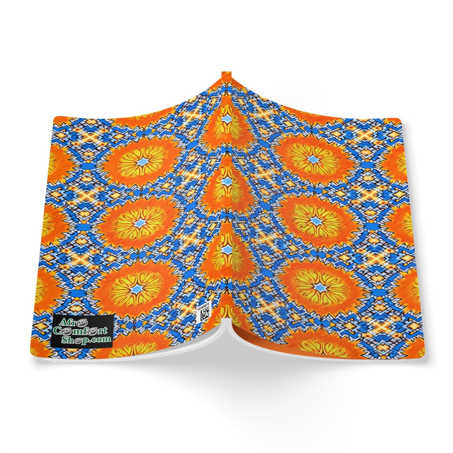 A colorful African-inspired journal displays a bold geometric pattern with orange and yellow sunbursts on a blue and white diamond background. The lower left patch reads “More Comfort For Them.” The book appears to float above a white surface.