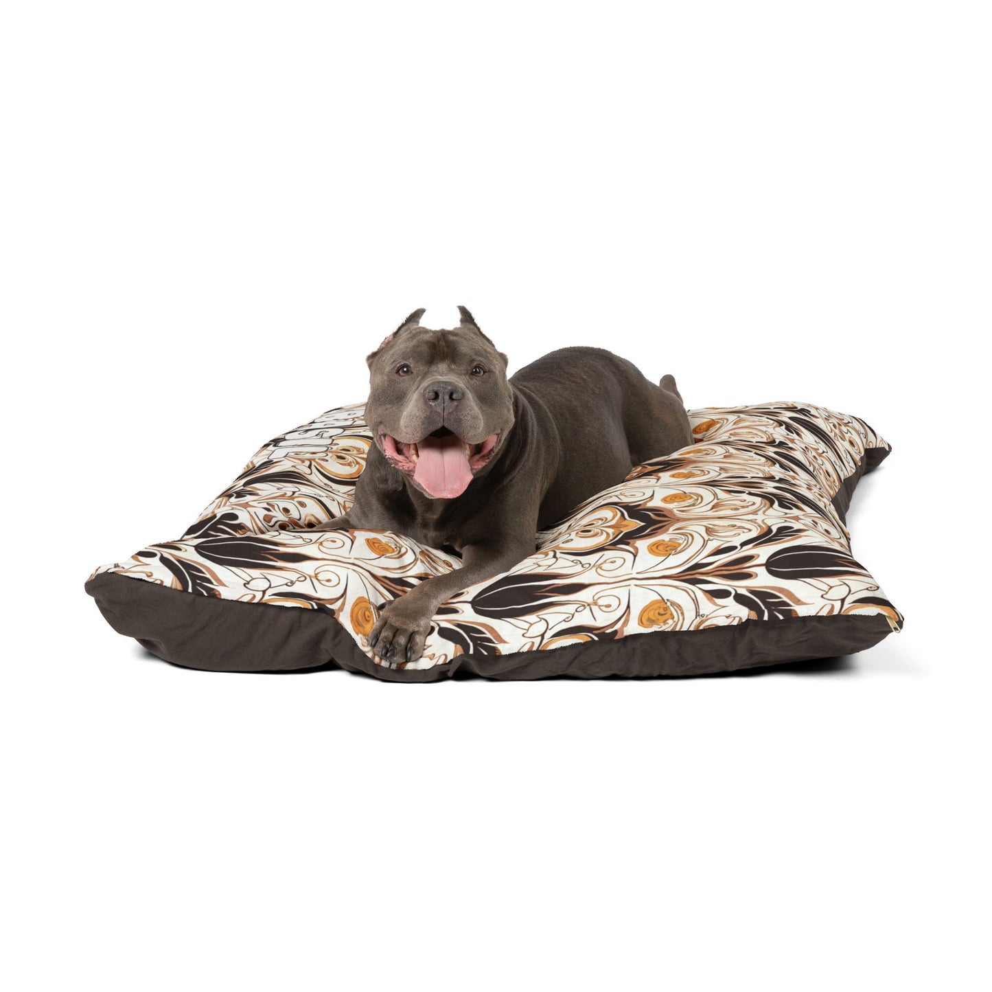 A gray dog with cropped ears and a pink tongue lies on a large, African-inspired patterned dog bed featuring brown, white, and orange floral designs. The dog appears relaxed and happy, facing forward with its mouth open and tongue out.