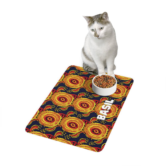 A white and gray cat sits on a mat with a bold, African-inspired pattern in red, yellow, and black. A white bowl of kibble rests on the mat beside the printed word BASIL in white, all set against a white backdrop.