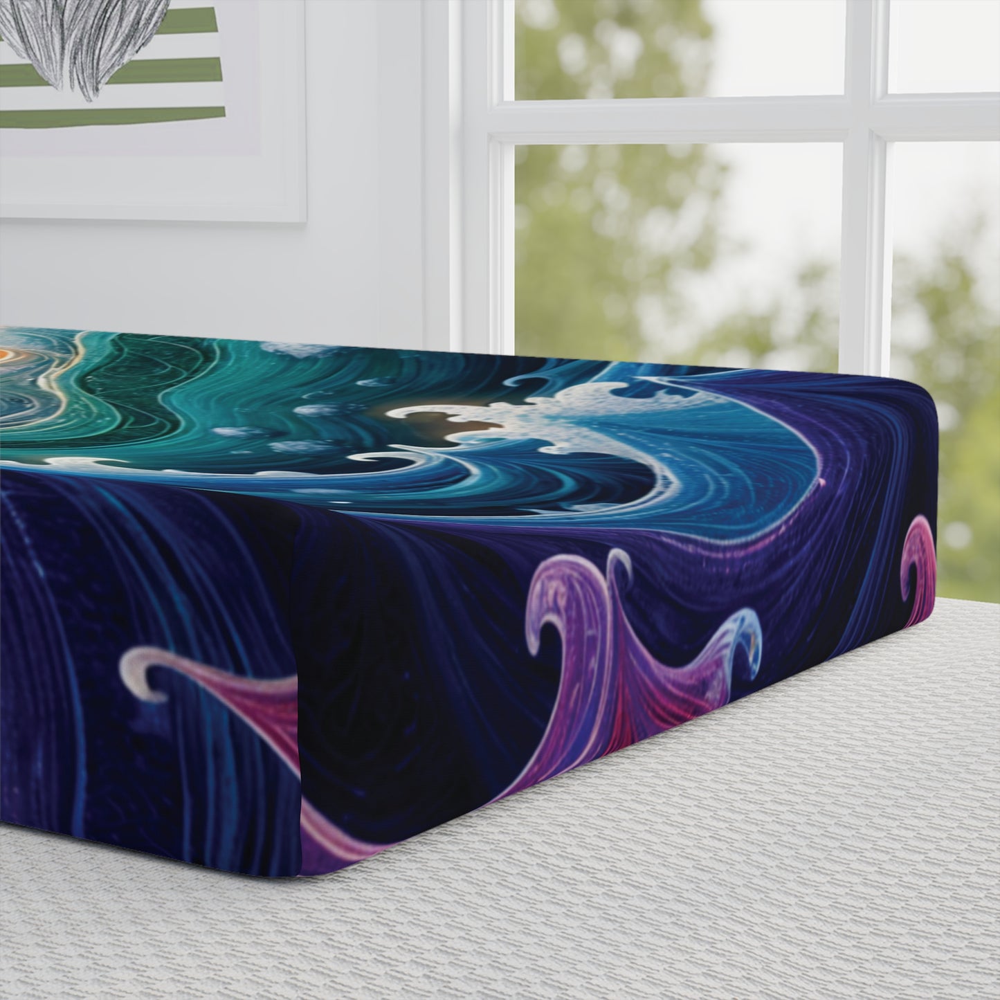 A close-up of a bed with a fitted sheet featuring a swirling abstract design in blue, teal, purple, and pink—reminiscent of African-inspired designs. The bed is by a large window with white framing, and part of a framed artwork is visible on the wall.