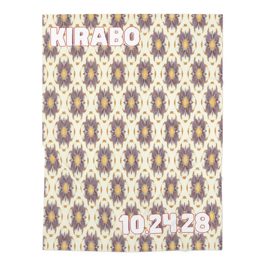 A cream personalized baby blanket with a repeating purple and gold floral pattern, inspired by African-inspired decor. KIRABO is in bold white and pink letters at the top left, with the date 10.24.28 in matching font at the bottom right.