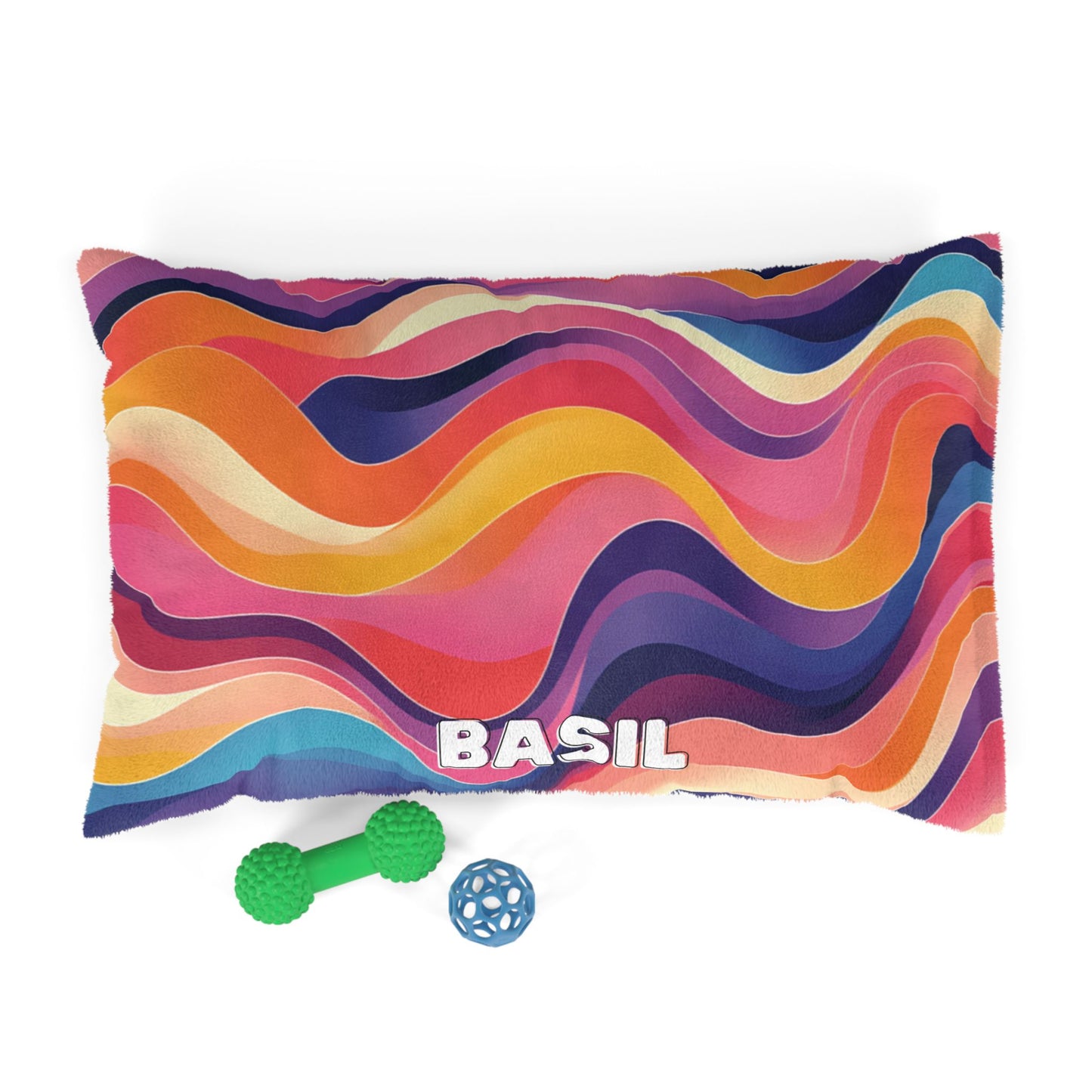 A colorful, African-inspired dog bed with wavy stripes in orange, pink, blue, and yellow tones. The name BASIL appears in bold white letters at the bottom. In front are a green bone-shaped toy and a small blue ball toy. White background.