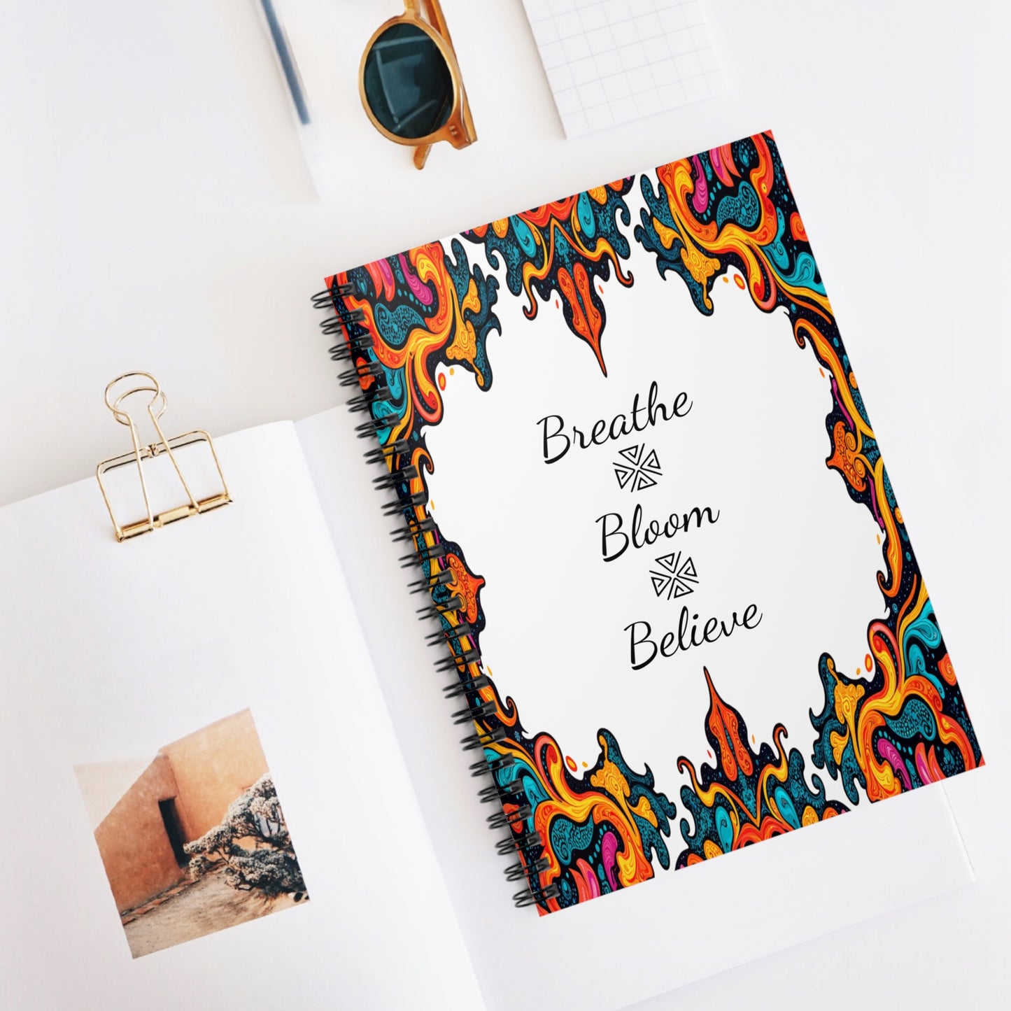 A spiral notebook with a colorful, ornate, flame-like border on its cover features culturally inspired designs. The center reads Breathe Bloom Believe in black script. It rests on a white desk with a photo book, sunglasses, binder clip, and grid paper nearby.