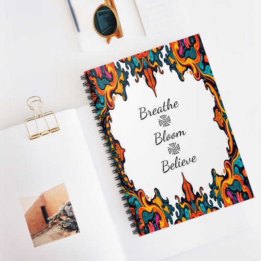 A spiral notebook with a colorful, ornate, flame-like border on its cover features culturally inspired designs. The center reads Breathe Bloom Believe in black script. It rests on a white desk with a photo book, sunglasses, binder clip, and grid paper nearby.