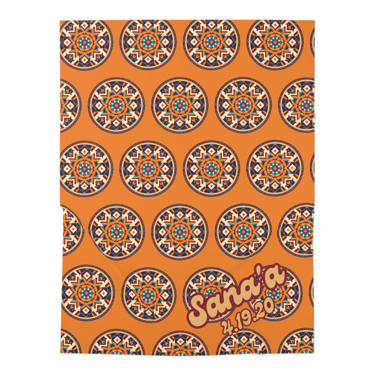 An orange baby swaddle blanket features a repeating pattern of circular geometric mandala designs in blue, white, and red. In the lower right corner, bold yellow text with a red outline reads “Sana’a 4.19.20.”, perfect for African-inspired decor.