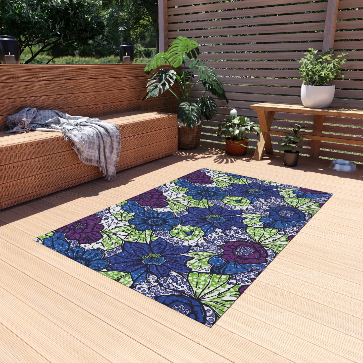 A colorful, weather-resistant outdoor rug with blue, purple, and green floral patterns lies on a wooden patio deck. Wicker furniture, striped throws, lush potted plants, and a bench complete the scene under bright natural daylight.