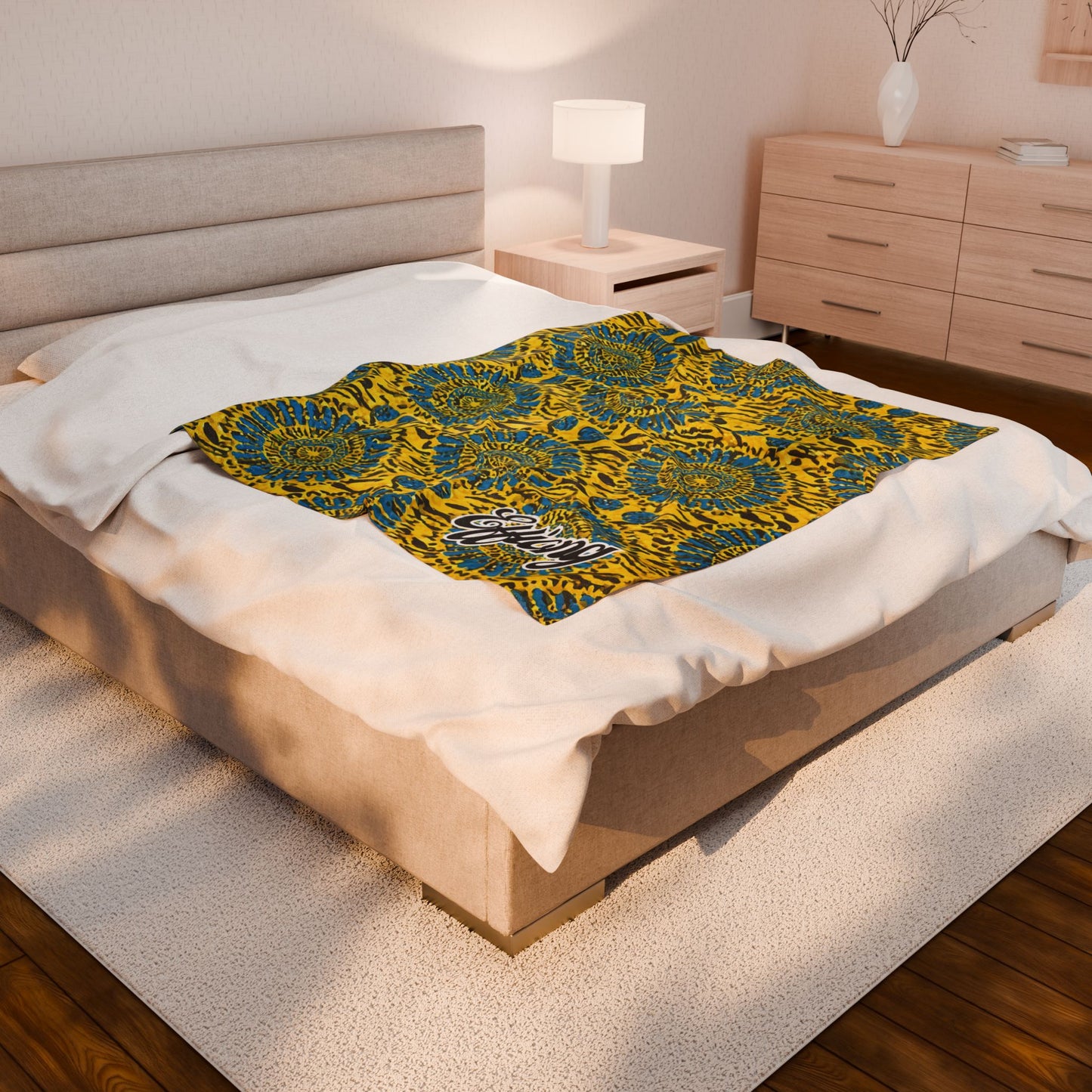 A modern bedroom with a beige upholstered bed, white bedding, and an African-inspired blanket in blue and yellow with large floral prints draped over the foot. The room features a wooden floor, white rug, nightstand, lamp, and dresser.