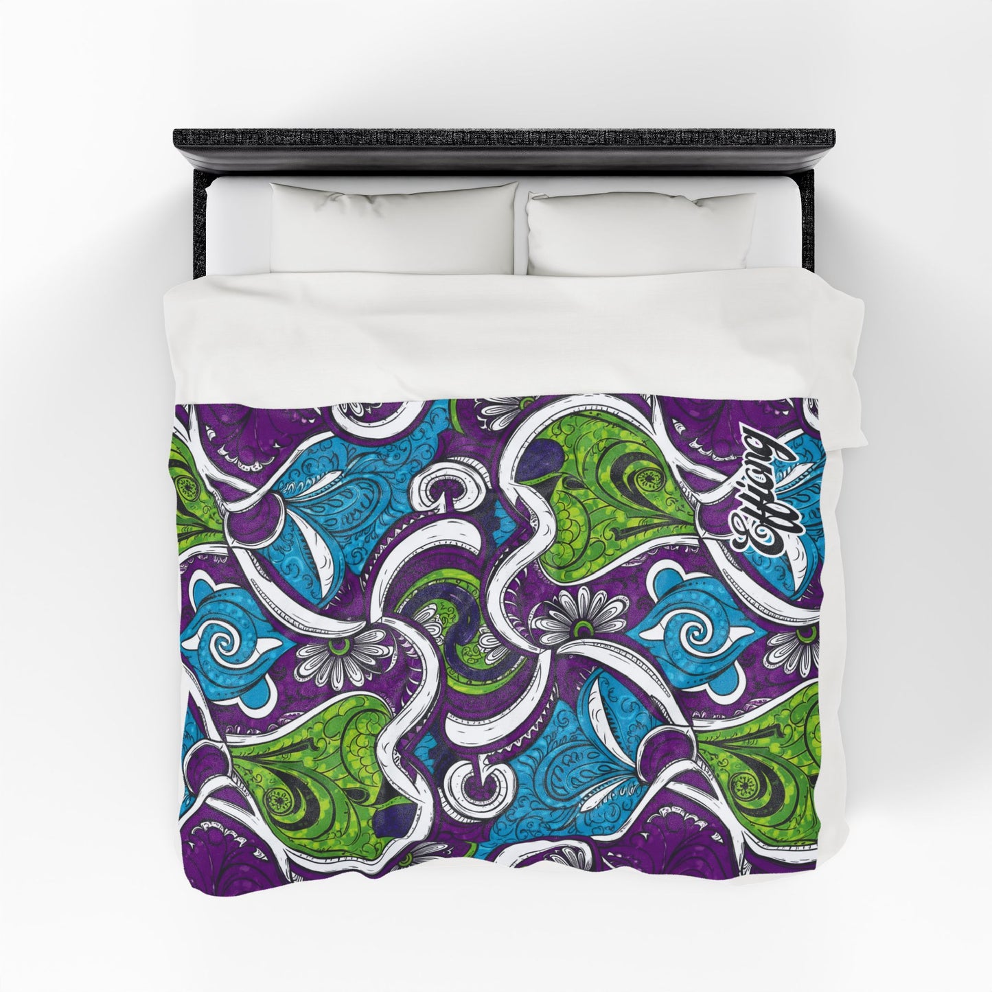 A bed with white pillows and an African-inspired blanket featuring a vibrant paisley pattern in purple, green, and blue, accented with swirling white and black lines. The corner displays stylized text reading “Artful Enigma.”.