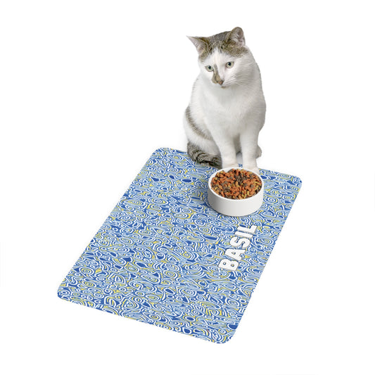 A white and gray cat sits on a blue African-inspired patterned mat with white swirls and the word fish printed on it. In front of the cat is a white bowl filled with dry cat food. The background is plain white.