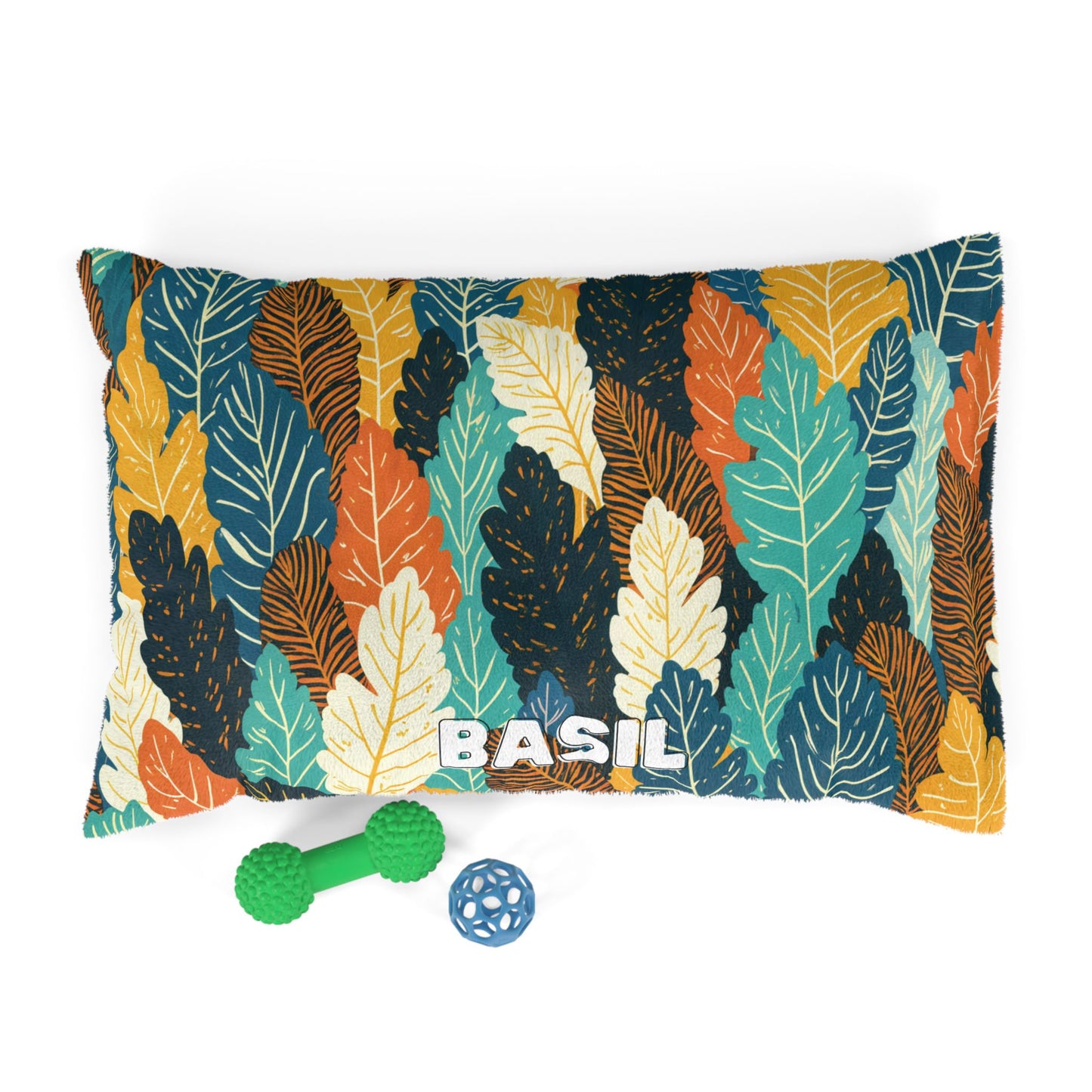 A rectangular pillow with a bold leaf pattern in teal, gold, orange, and white offers luxurious comfort. The name BASIL is printed in large white letters at the bottom center. In front are two green and blue dog toys on a white background.