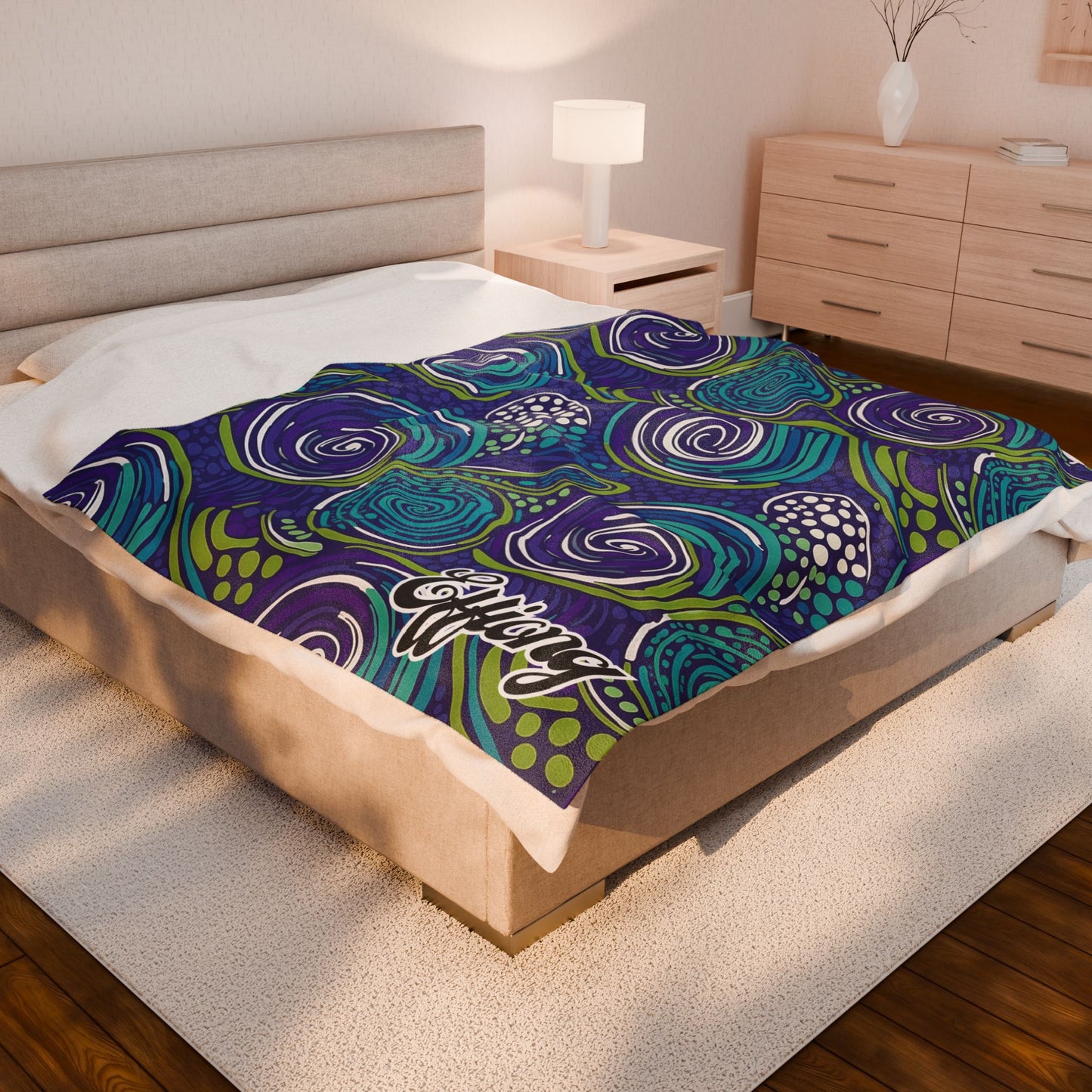 A bedroom with a beige upholstered bed, white bedding, and a unique African-inspired personalized blanket featuring blue, green, and purple abstract swirl patterns. A nightstand with lamp, wooden dresser, vase with branches, and beige rug complete the look.