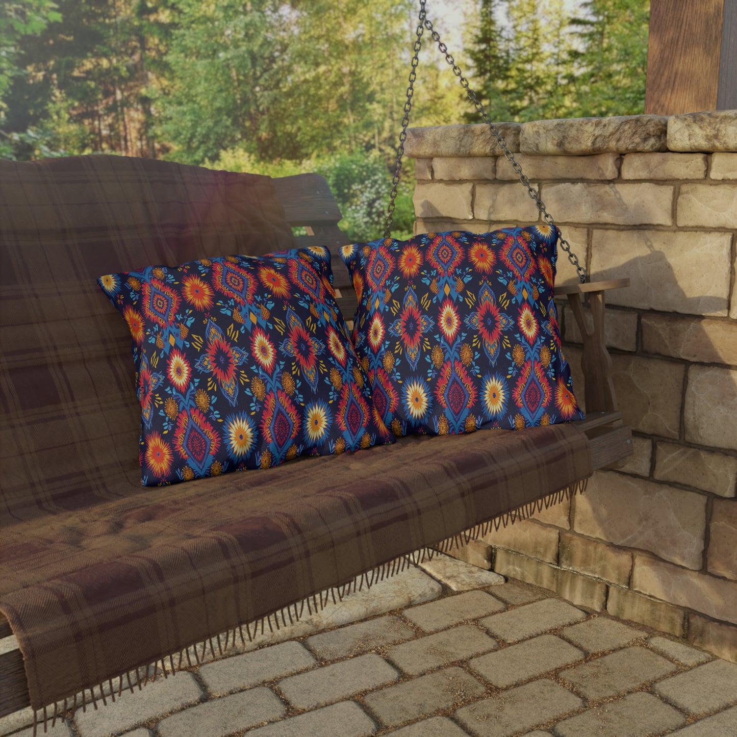 A wooden porch swing with UV-resistant outdoor pillows and a plaid blanket sits next to a stone wall, overlooking a patio and green, sunlit trees in the background.