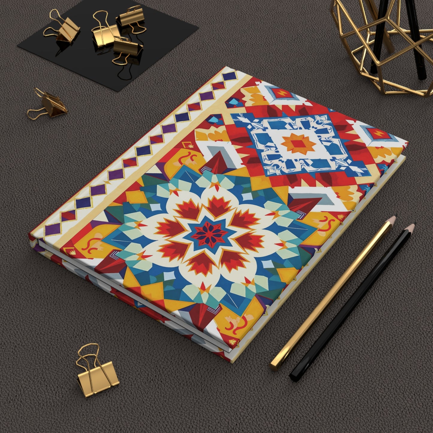 A closed hardcover journal notebook with a vibrant, African-inspired design and floral pattern rests on a textured brown surface. Nearby are black and gold pencils, gold binder clips, and a decorative geometric metal object—all neatly arranged.