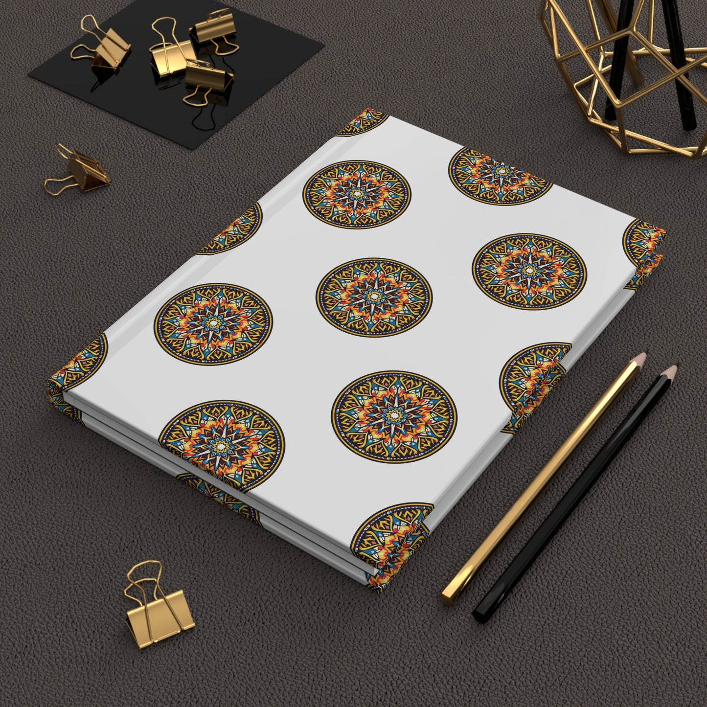 A closed African-inspired hardcover journal with a white cover featuring colorful, intricate circular mandala patterns lies on a dark textured surface. Nearby are gold and black pencils, gold binder clips, and a geometric gold wire decor item.