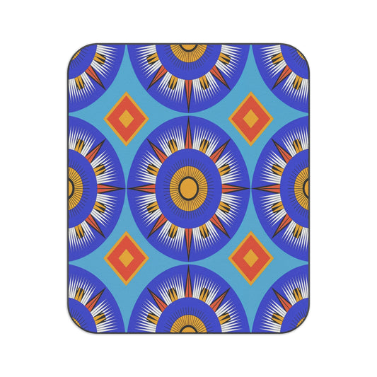 A rectangular mat with rounded corners features a bold, symmetrical pattern of blue, yellow, and white sunburst circles and red-orange diamonds on a light blue background. This African-inspired picnic blanket is geometric and vibrant, creating a lively visual effect.