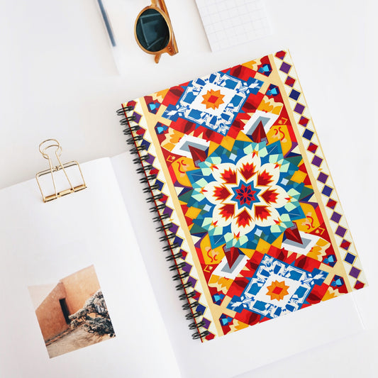An African-inspired spiral notebook with a colorful, geometric, mandala-like cover design sits on a white desk. Nearby are a binder clip, sunglasses, a grid notepad, and an open book displaying a photo of a wall and stone steps.