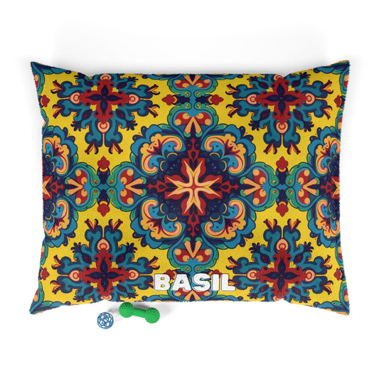 A rectangular custom pet pillow with a vibrant, ornate yellow, blue, and red floral pattern. The name BASIL is printed in white near the bottom. Below the stylish dog bed are a small green bone-shaped toy and a blue and white ball.