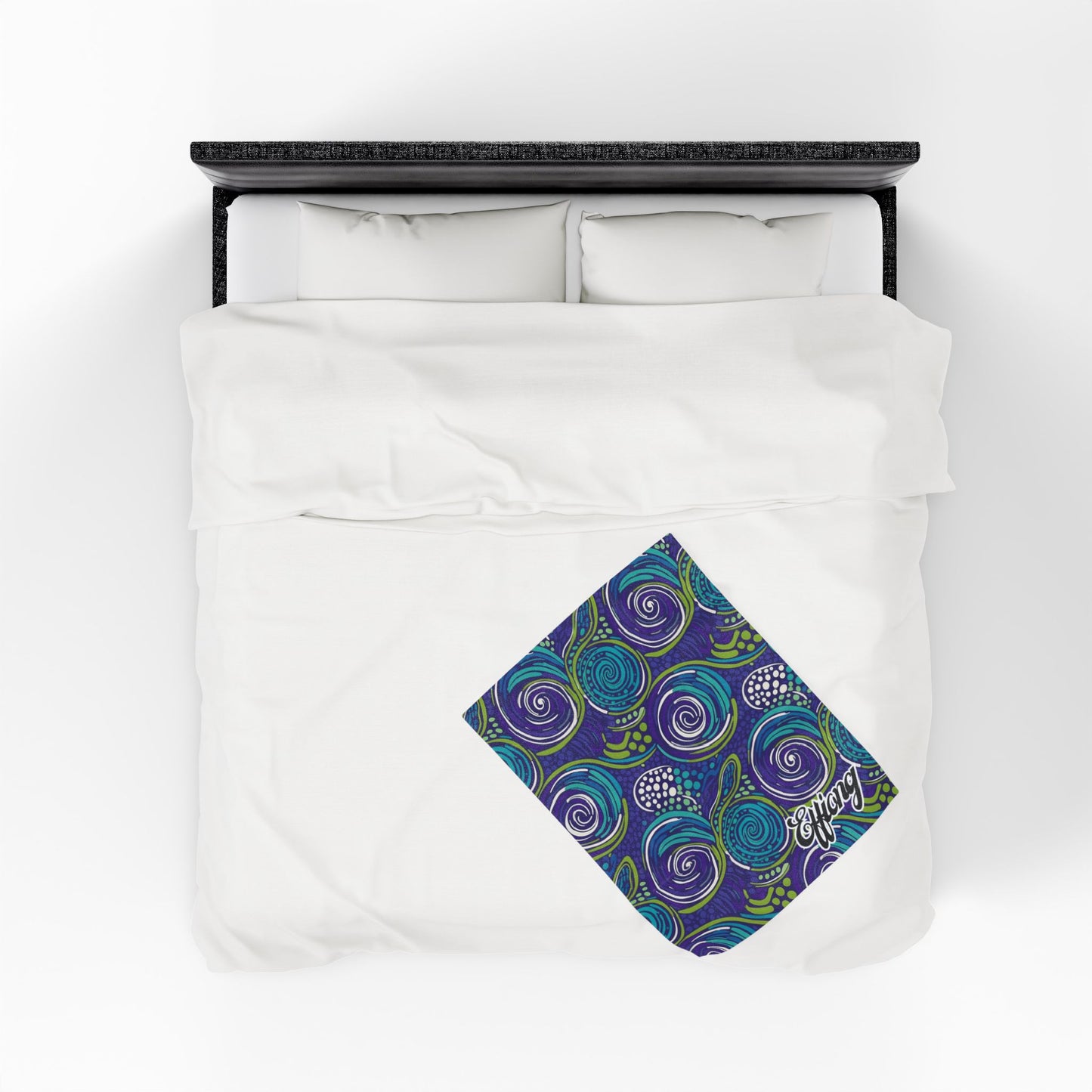 A white bed with two pillows seen from above. At the foot lies a personalized velveteen blanket featuring vibrant blue, purple, and green abstract swirls and leaves, with “energy” written in cursive in one corner.