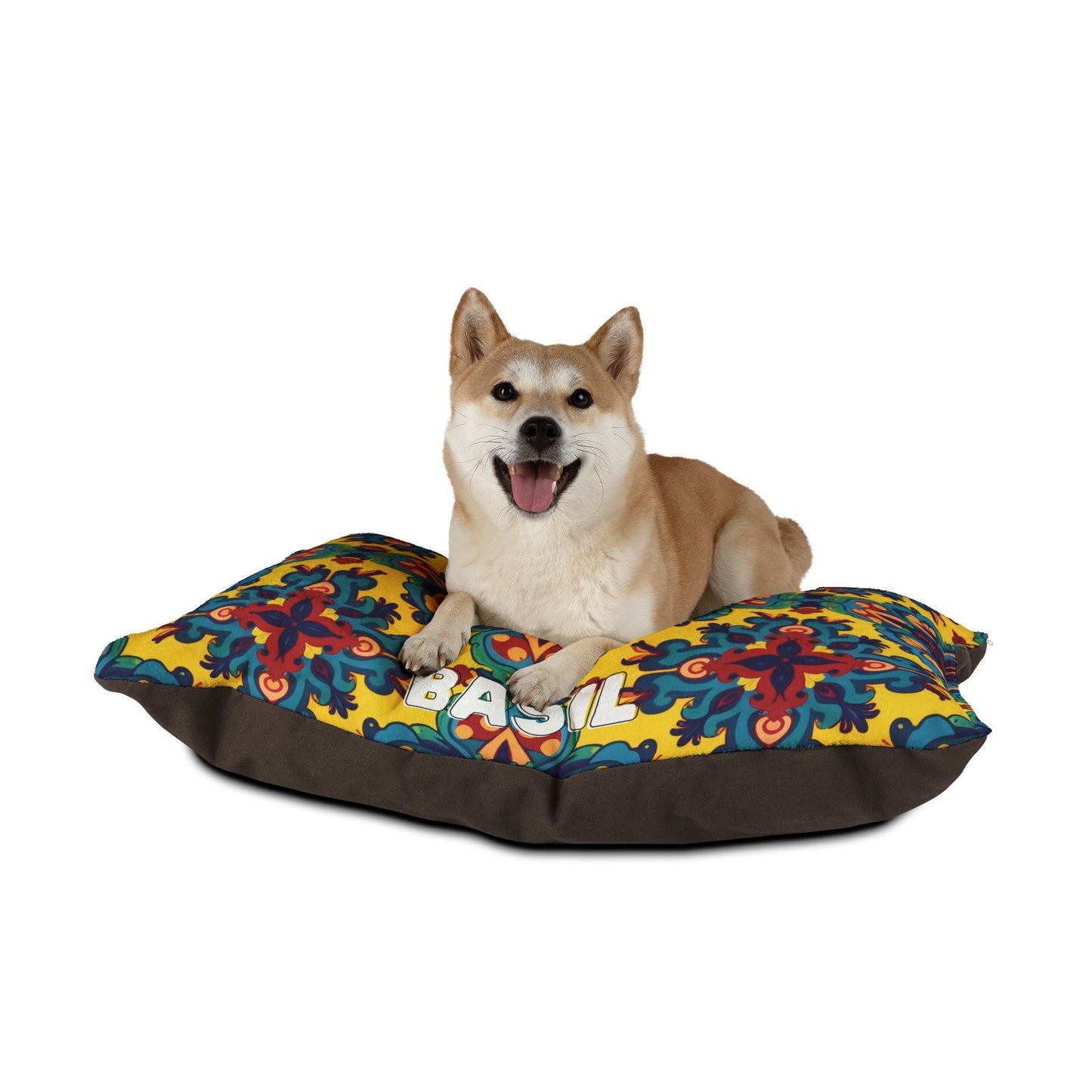 A Shiba Inu dog sits on a colorful, African inspired pet bed with blue, red, and yellow designs and the word BASIL visible. The happy dog looks forward with its mouth open against a plain white background.