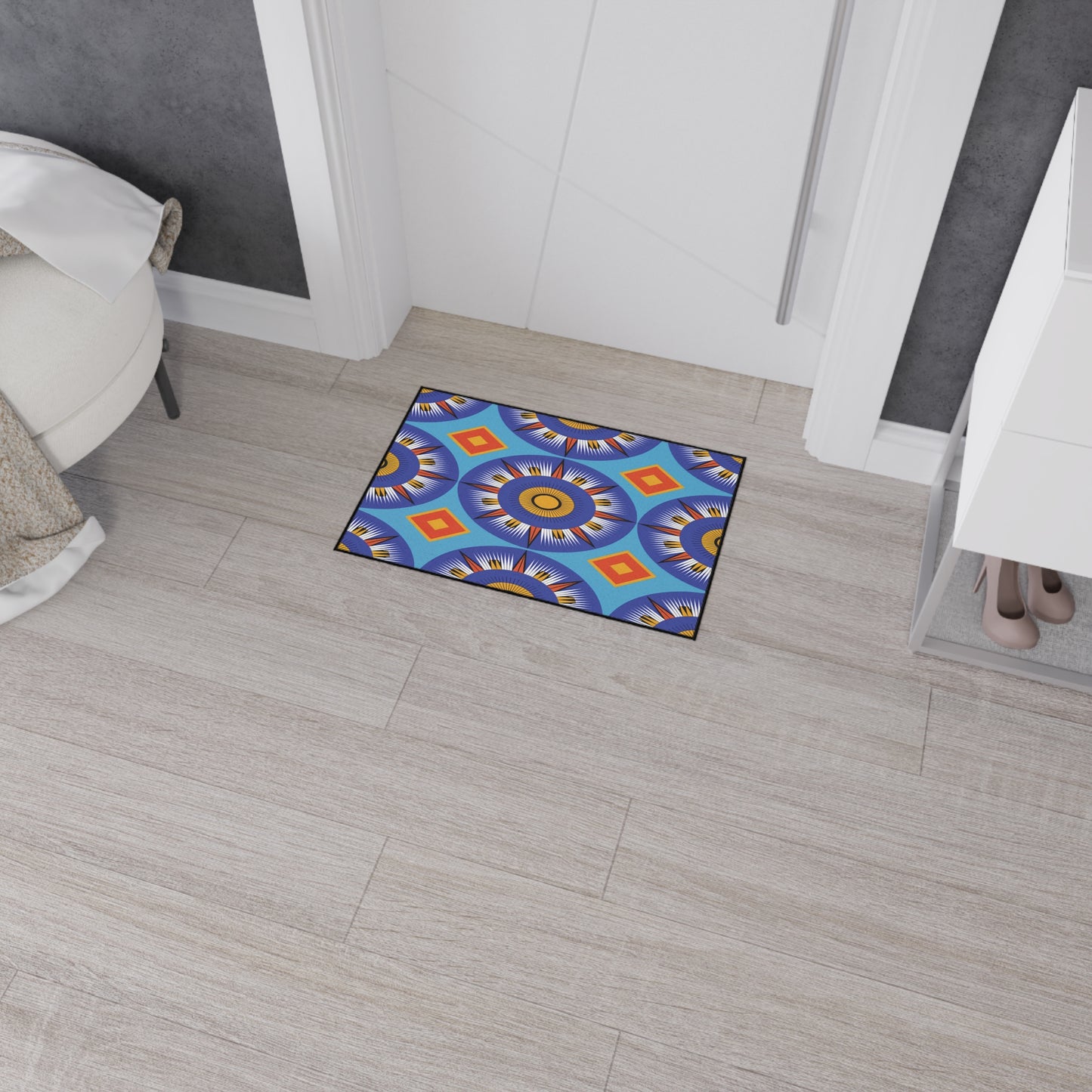 A small doormat with a vibrant, African-inspired geometric pattern in blue, yellow, orange, red, and purple sits on light wood floor tiles near a white door. It features a non-slip rubber backing for safety and rests beside a cabinet and soft round beige seat.