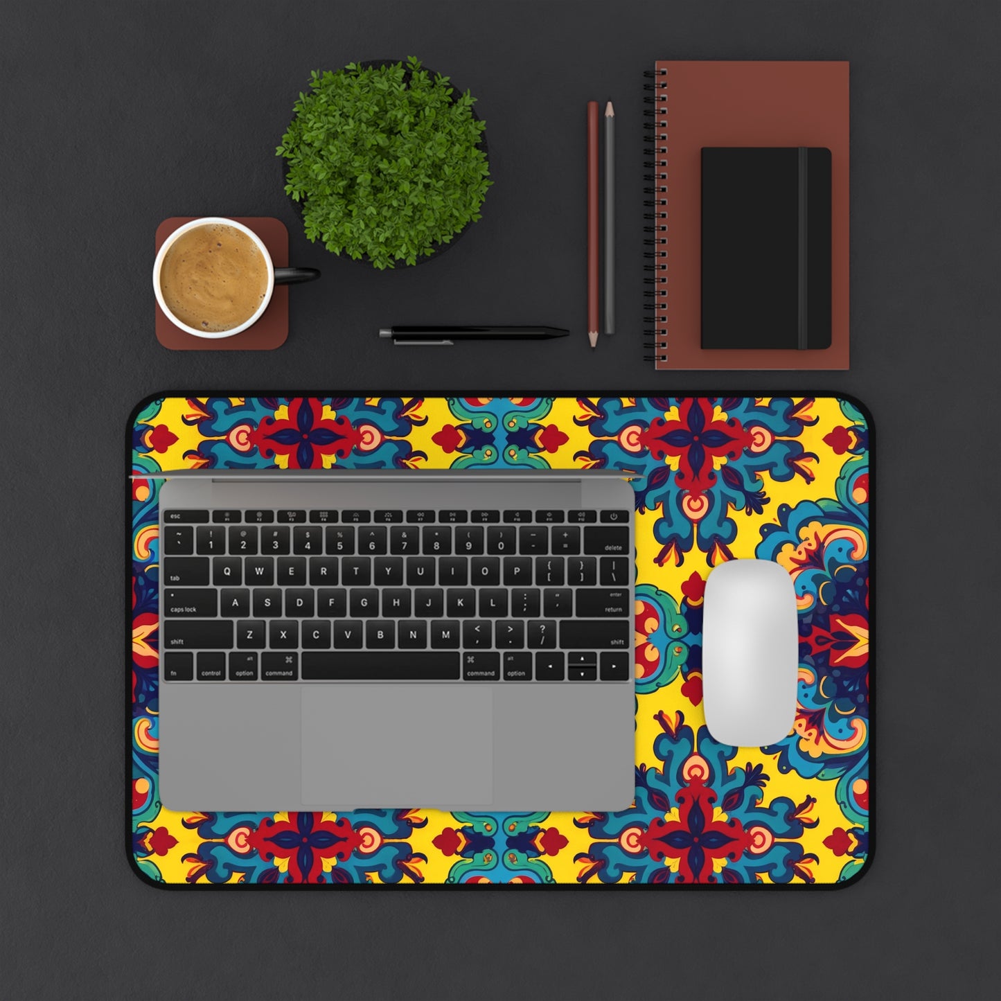 A laptop and white mouse sit on a colorful neoprene desk mat with red, blue, and yellow designs. Nearby are a brown notebook, writing tools, a small green potted plant, and coffee on a brown saucer atop the dark surface.