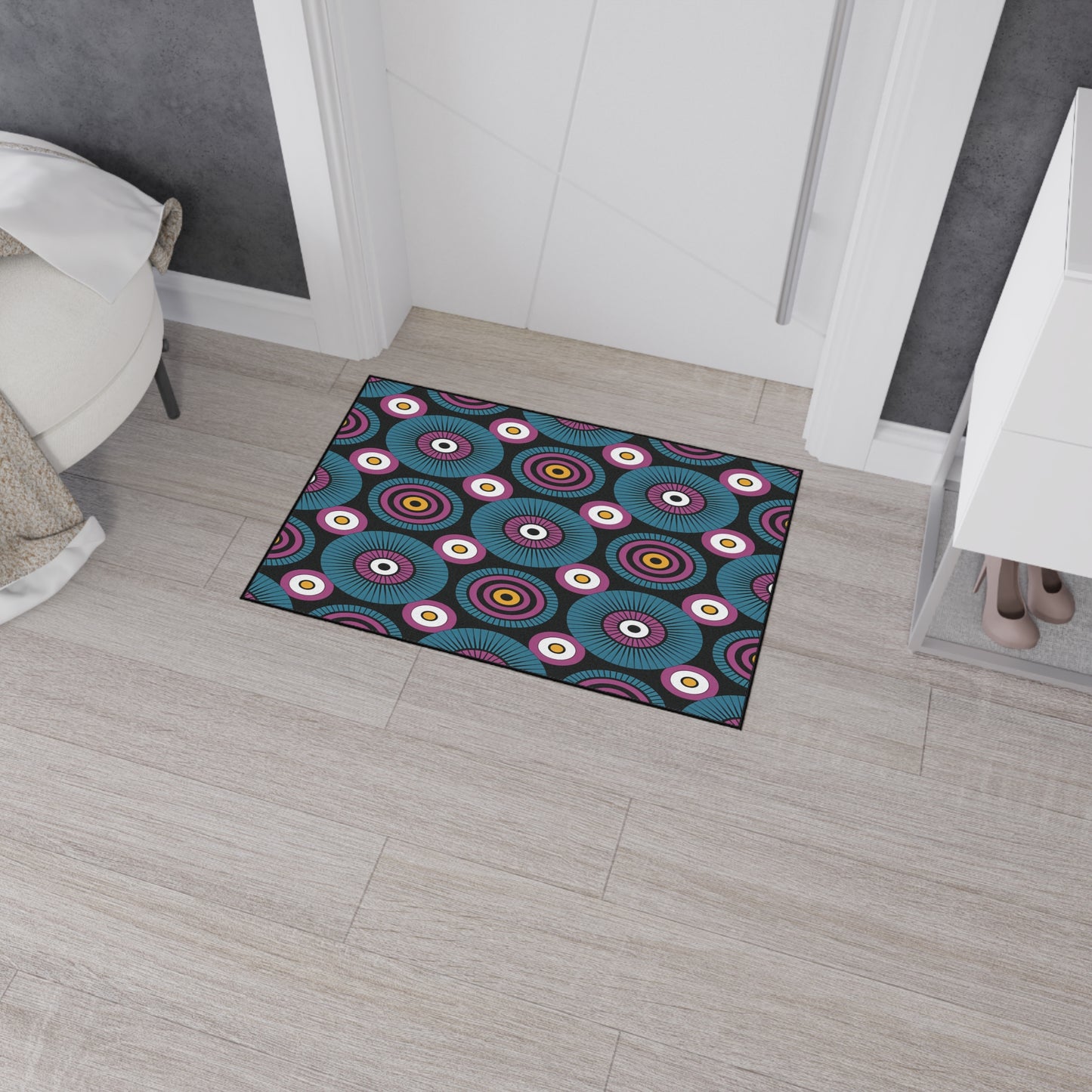 An African-inspired, non-slip doormat with a colorful circular pattern featuring concentric rings and eye-like designs lies on a light wood floor near a white door. Nearby are a white cabinet with shoes inside and part of a round chair.