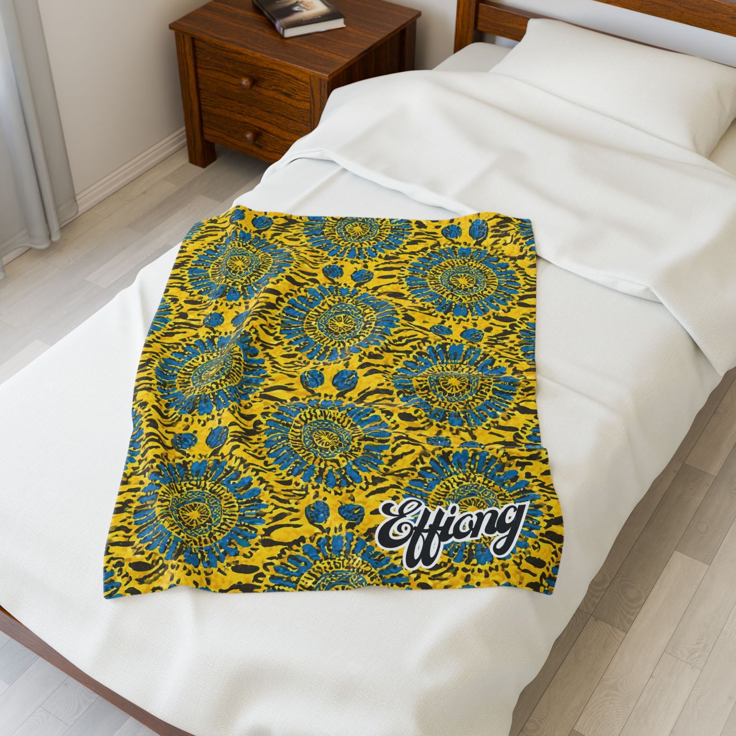 A vibrant yellow and blue African-inspired blanket with a bold, circular floral design and “Effiong” in white script lies neatly over a white blanket on a made bed. Beside the bed, a wooden nightstand holds two books.