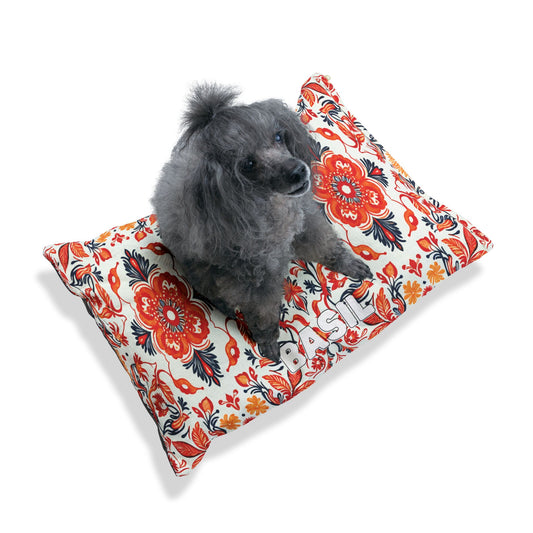 A small, curly-haired gray dog sits on a colorful, African-inspired pillow decorated with red and orange floral patterns and the word BASIL in bold white letters. The plain white background keeps the focus on the dog and vibrant pillow.